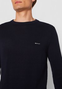 Pulover tricotat de culoare bleu marin, cu guler rotund și finisaj texturat, având un mic logo GANT alb pe piept.