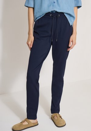 Femme portant un pantalon bleu marine à cordon, une chemise bleu clair boutonnée et des sandales beiges à enfiler avec détail de boucle.