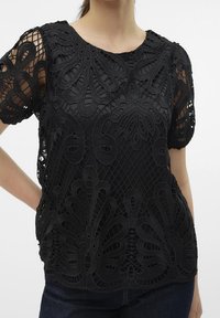 Haut en dentelle noire avec un design floral et des éléments transparents, doté d'un col rond. Motif en maille texturée avec des manches courtes et une coupeLoose.