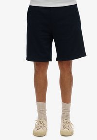 Pantaloni corti blu navy realizzati in misto cotone, con vita elasticizzata, tasche laterali e orli cuciti. Indossati con calzini e scarpe di colore chiaro.
