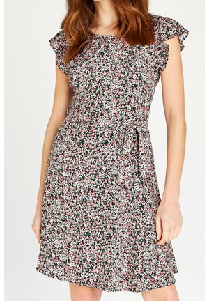 Kobieta ubrana w sukienkę bez rękawów w kwiatowy wzór z falbankami na ramionach i przewiązanym paskiem w talii na jednolitym tle.