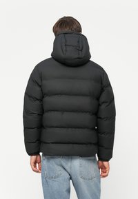 Homme portant une veste doudoune noire avec capuche et un jean bleu clair, vu de dos sur un fond blanc uni.