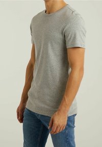 Homme portant un t-shirt uni gris clair à manches courtes et un jean bleu, debout avec les bras détendus sur un fond neutre.