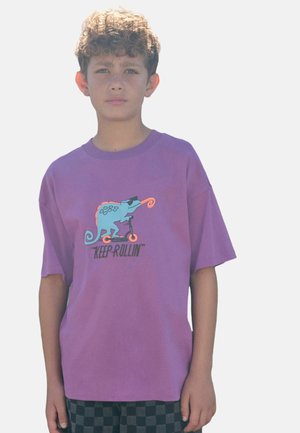 T-shirt viola a maniche corte con un camaleonte dei cartoni animati che guida uno scooter, con accenti colorati e il testo "CONTINUA A RULLARE".
