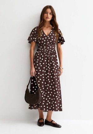 Vrouw die een bruine midi-jurk met witte stippen draagt, een donkerbruine handtas vasthoudt en tegen een witte muur staat.