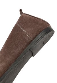 Bottine en daim marron avec une semelle en caoutchouc noir texturé. Elle présente des surpiqûres blanches contrastantes et une languette à l'arrière pour un enfilage facile.
