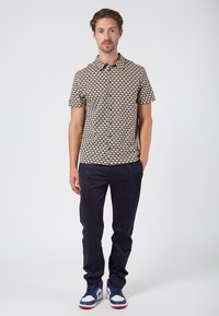 Korte mouwen shirt met een crème basis, voorzien van een geometrisch blauw en bruin patroon. Gepaard met donkerblauwe broek en blauwe sneakers.