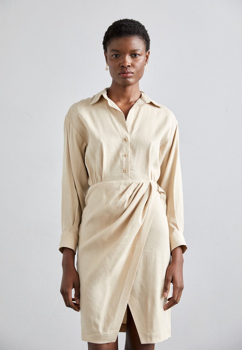 Vestido camisa beige con mangas largas, botonera en la parte delantera y detalle envolvente fruncido en la cintura, con una abertura lateral y textura suave.