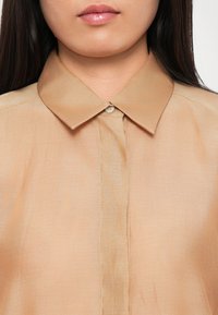 Beige knappskjorte med klassisk krage, laget av et glatt, lett stoff med en subtil glans. Enkel fremre placket med en enkelt knapp.