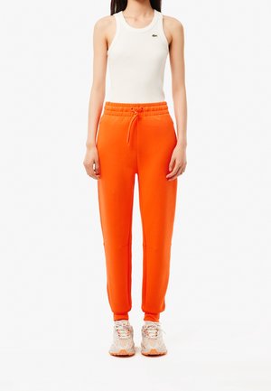 Kvinde iført en hvid ærmeløs ribbed top og lyse orange træningsbukser med snøre, samt beige sneakers, står mod en almindelig hvid baggrund.
