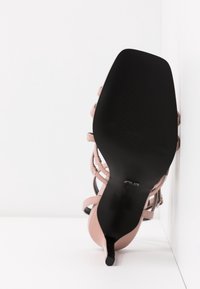 Sandale à talons rose avec des brides noires, présentant une silhouette courbée, une texture lisse et un bout pointu. Semelle en caoutchouc noire.