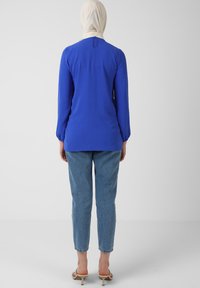 Top azzurro a maniche lunghe con polsini arricciati, chiusura a bottoni al collo, abbinato a jeans in denim chiaro e sandali beige.