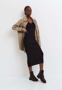 Robe en tricot côtelé noire portée avec une veste en simili cuir tan. Le mannequin porte des bottines noires avec des rayures rouges et une fermeture éclair.