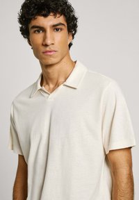 Cream short-sleeve polo shirt with a v-neck design, featuring a textured fabric and subtle stitching details. Soft, lightweight material.