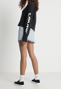 Chemise noire courte à manches longues avec logo "Levi's" blanc, associée à un short en jean bleu clair déchiré et des baskets noires avec des chaussettes blanches.