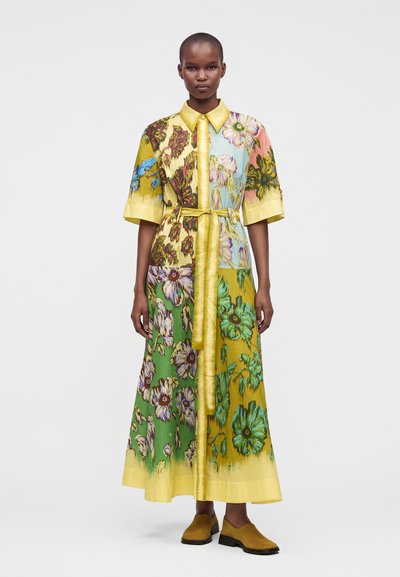Femme debout portant une longue robe fleurie multicolore avec un col jaune, une taille ceinturée, des manches courtes et des chaussures slip-on moutarde.