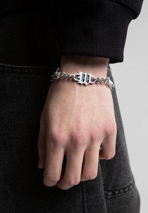 Ασημένιο βραχιόλι αλυσίδας με λεπτομέρειες από συρματόπλεγμα και γοτθικό γράμμα "W", φορεμένο στον καρπό ενός ατόμου πάνω από μαύρο μανίκι και σκούρο παντελόνι.