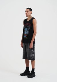 Camiseta sin mangas negra con un diseño gráfico colorido, combinada con unos pantalones cortos de mezclilla grises hasta la rodilla con un estampado. Unos zapatos negros completan el conjunto.