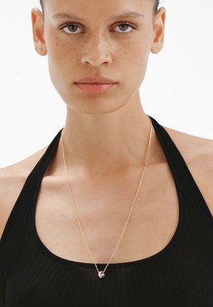 Collar de cadena de oro con un colgante curvado. La modelo lleva una camiseta negra de canalé, mostrando la simplicidad y el brillo de la cadena.