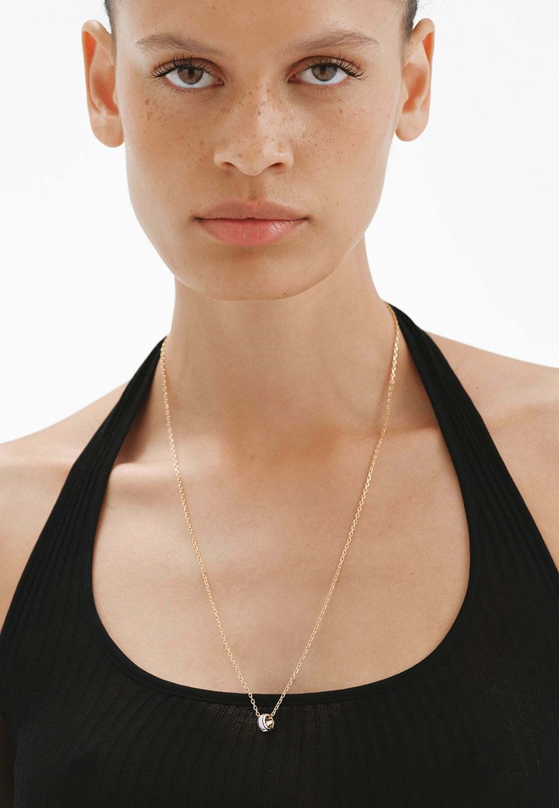 Collar de cadena de oro con un colgante curvado. La modelo lleva una camiseta negra de canalé, mostrando la simplicidad y el brillo de la cadena.