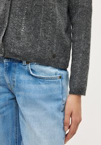 Grijze gebreide cardigan met een gestructureerd patroon, voorzien van knoopsluitingen en pailletten. Gecombineerd met lichtblauwe spijkerbroek.