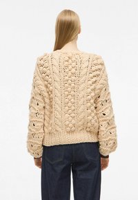 Gruba be�żowa sweter z dzianiny, z teksturowanymi wzorami, kulkami i dopasowanym pasem. Długie rękawy z ażurowymi detalami i ściągaczami.
