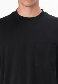T-shirt en coton noir avec un col rond, doté d'une poche poitrine et d'une broderie de logo discrète. Texture lisse et coupe décontractée.