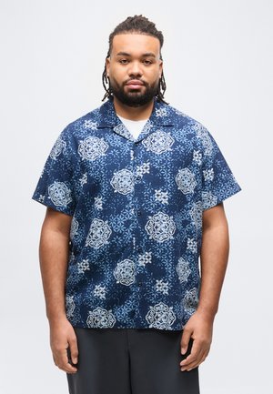 Man with short dreadlocks and beard wearing a blue patterned short-sleeve button-up shirt and dark pants, standing against a plain background.