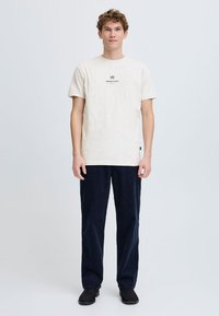 Camiseta de algodón beige de mangas cortas y cuello redondo, con un pequeño logo negro en la parte delantera, combinada con pantalones de pana en tono azul marino oscuro.