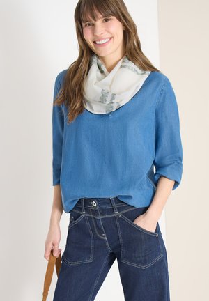 Femme souriante, portant un haut à manches longues bleu, une écharpe blanche à motifs, un jean bleu foncé, et tenant une sangle de sac à main beige dans une main.