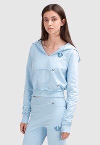 Jasnoniebieska krótka bluza z szerokim kołnierzem, kieszenią z przodu i dekoracyjnymi guzikami. Posiada postrzępione wykończenie i haftowane logo na klatce piersiowej.