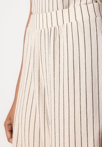 Lyse fargede stripete bukser med vertikale svarte striper, med teksturert stoff og plisserte detaljer i midjen.