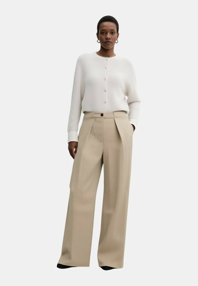 Donna in piedi, indossa una camicetta bianca a maniche lunghe con bottoni e pantaloni beige a gamba larga, con una mano in tasca, sfondo neutro.