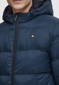 Tamsiai mėlynas pūkų paltas su užtrauktuku, gobtuvu ir juodomis detalėmis. Yra siuvinėta struktūra ir mažas logotipas ant krūtinės.