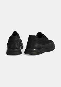 Black sneakers featuring a textured leather upper, lace closure, and a chunky rubber sole with a smooth finish. Rear view shows a rounded silhouette.