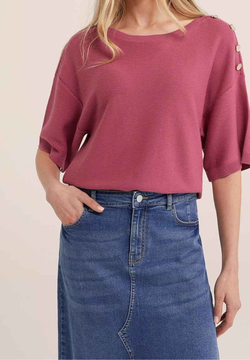 Femme portant un haut en maille rose avec des détails de boutons sur la manche et une jupe en jean bleue taille haute avec des poches.