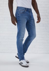 Uske slim-fit plavi traperice s umjerenim pojasom, pet džepova i suptilnim izbljedjelim detaljima, uparenim s bijelim i sivim tenisicama.