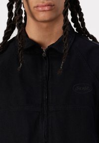 Svart, zip-up jakke med krage, med et subtilt logo-patch og teksturert stoff. Designet inkluderer raglanermer og flate sømmer.