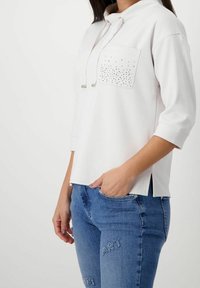 Felpa bianca con cappuccio e tasca frontale caratterizzata da dettagli con borchie. Leggera miscela di cotone con maniche a tre quarti, abbinata a jeans blu.