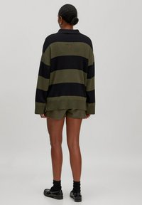 Übergroßer schwarz-oliv gestreifter Pullover mit Kragen, kombiniert mit olivfarbenen Shorts. Das Material scheint gestrickt zu sein und verfügt über seitliche Schlitze.
