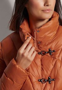 Oranje pufferjack met een hoge kraag, voorzien van zwarte hardware toggles, ritssluiting en een gequilte patroon. Glad, glanzend textuur.