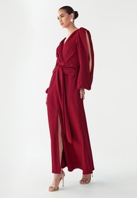 Pikk, punane mähkkleit, millel on pikad varrukad, küljetagused ja vööga seotud talje. Sile kangas voolava disainiga, kombineeritud rihmadega metallist kontsadega.