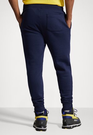 Personne portant un pantalon de jogging bleu marine et des chaussures de randonnée jaunes, bleues et noires, debout les mains dans les poches.