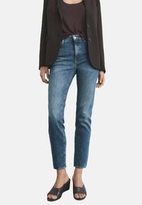 Höga blå denimjeans med slim fit, med fem fickor och knappstängning, i kombination med en svart kavaj och öppna klacksandaler.