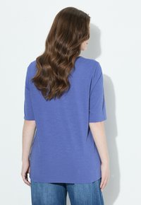 Mujer con cabello largo y ondulado de color castaño, vestida con una camisa azul de manga corta y holgada, y jeans azules, de pie frente a una pared blanca.