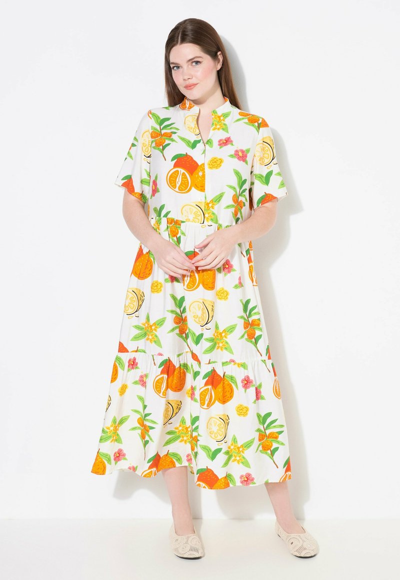 Vestido maxi blanco con patrones de frutas cítricas en naranja y amarillo, y flores; mangas cortas, cierre de botones en la parte delantera y diseño en capas. Zapatos de ganchillo beige.