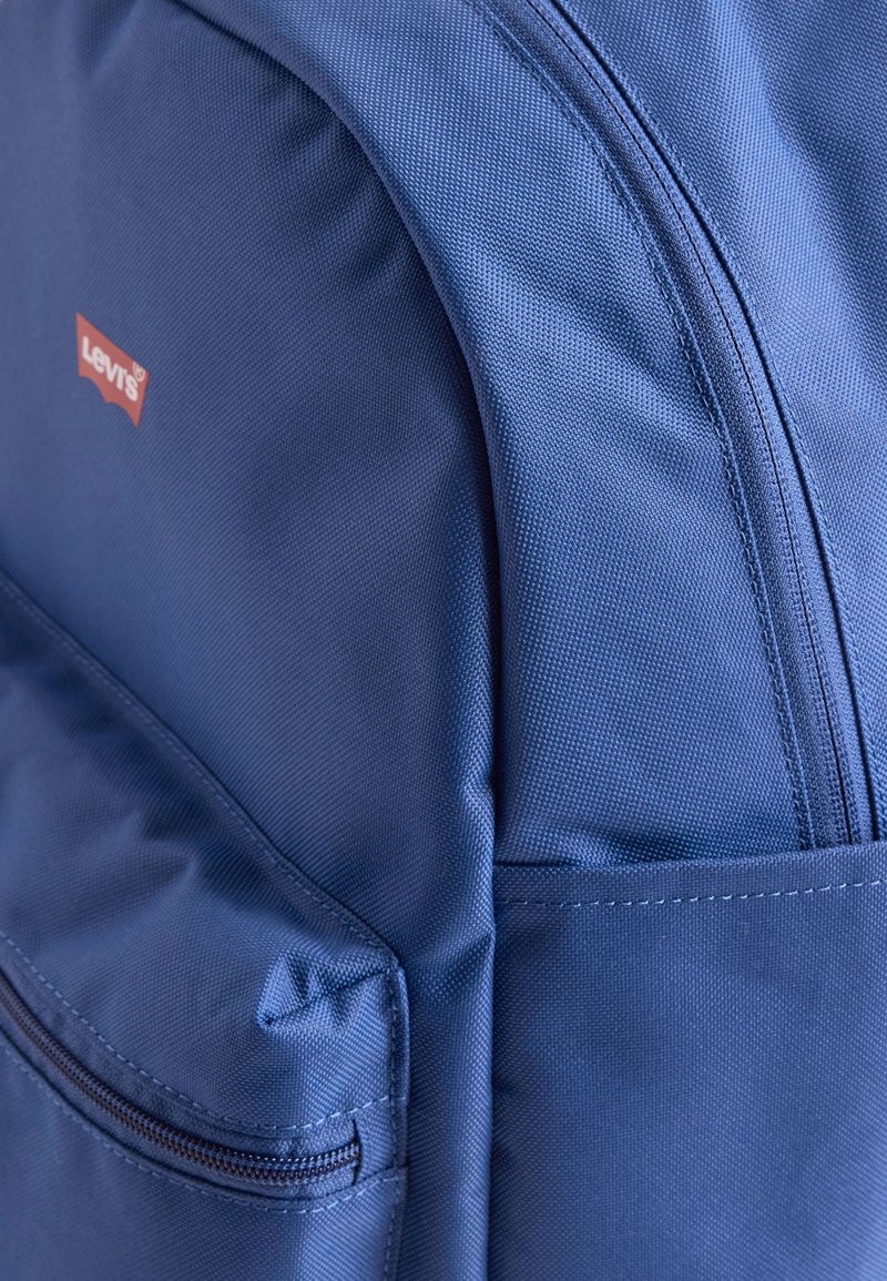 Mochila de tela azul con un bolsillo frontal con cremallera y un parche del logo de Levi's en rojo en la parte superior izquierda.