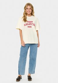 Krémové oversized tričko s červeným grafickým textem "PARIS COLETTE CLUB", doplněné světle modrými širokými džínami a hnědými nazouvacími botami.