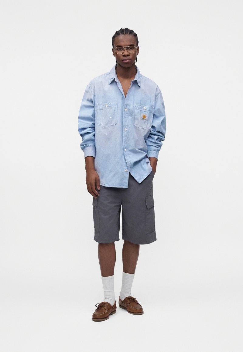 Giovane uomo con occhiali, camicia azzurro chiaro a maniche lunghe, pantaloncini cargo grigi, calzini bianchi e scarpe di pelle marroni, in piedi su sfondo bianco.