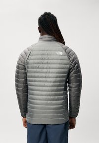 Grijze geïsoleerde jas met verticale quiltstructuur, hoge kraag en het logo van The North Face op de achterkant. Gladde textuur met een lichte glans.